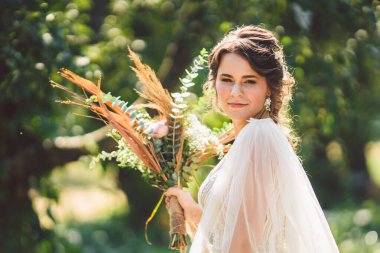 Çiçekli güzel gelin orman arka planında duruyor. Kırsal tarzda. Dışarıda zarif elbiseler içinde güzel bir gelin. Güneşli, ağaçlık alanda parkta genç bir gelinin portresi.