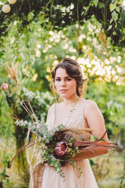 Çiçekli güzel gelin orman arka planında duruyor. Kırsal tarzda. Dışarıda zarif elbiseler içinde güzel bir gelin. Güneşli, ağaçlık alanda parkta genç bir gelinin portresi.
