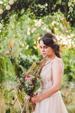 Çiçekli güzel gelin orman arka planında duruyor. Kırsal tarzda. Dışarıda zarif elbiseler içinde güzel bir gelin. Güneşli, ağaçlık alanda parkta genç bir gelinin portresi.