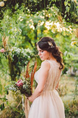Çiçekli güzel gelin orman arka planında duruyor. Kırsal tarzda. Dışarıda zarif elbiseler içinde güzel bir gelin. Güneşli, ağaçlık alanda parkta genç bir gelinin portresi.