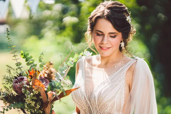 Schöne Braut mit Blumenstrauß steht auf Waldhintergrund. rustikaler Stil. schöne Braut in zartem Kleid im Freien. Nahaufnahme Porträt der jungen Braut im Park bei sonnigem Wetter Waldgebiet — Stockfoto