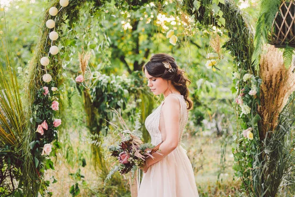 花束の花を持つ美しい花嫁は森の背景に立っています。素朴なスタイルだ美しい花嫁は繊細なドレスを屋外で。天気の良い日に公園で若い花嫁の肖像画を閉じる — ストック写真