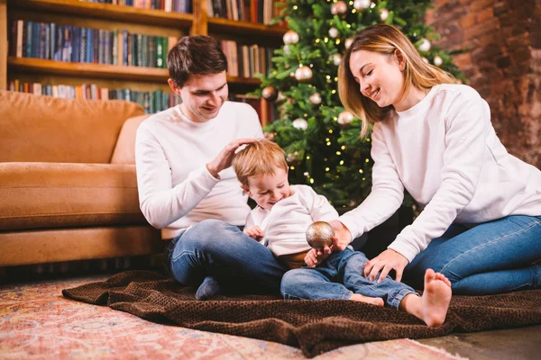 Concepto de Navidad familiar. La encantadora familia se sienta cerca del árbol de Navidad en la noche de invierno. Familia feliz con hijo en el suelo cerca del sofá En casa. Familia en casa en Año Nuevo. familia disfrutar de vacaciones de año nuevo — Foto de Stock