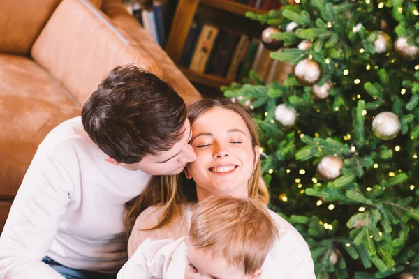 Concepto de Navidad familiar. La encantadora familia se sienta cerca del árbol de Navidad en la noche de invierno. Familia feliz con hijo en el suelo cerca del sofá En casa. Familia en casa en Año Nuevo. familia disfrutar de vacaciones de año nuevo — Foto de Stock