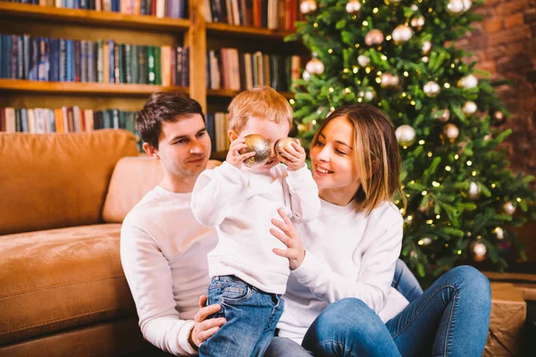 Conceito de Natal de Família. Linda família senta-se perto da árvore de Natal na noite de inverno. Família feliz com filho no chão perto do sofá em casa. Família em casa no Ano Novo. família desfrutar de feriados de ano novo — Fotografia de Stock