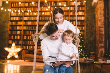 Aile birlikteliği. Anne, baba ve oğul salıncakta birlikte kitap okuyorlar. Aile ve Ebeveynlik kavramı. Mutlu aile çatı katında otururken çocuk kitapları okuyor.