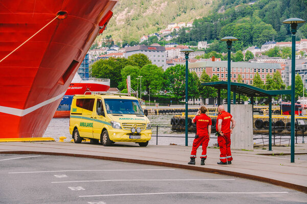 Two Norwegian paramedics in red uniforms are resting near an ambulance parked in a port near a large ship. Theme healthcare and medicine in Norway, Bergen July 28, 2019