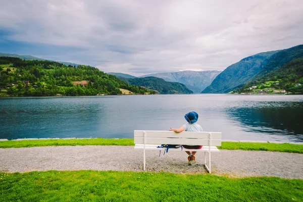 在挪威乌尔维克，一个女人坐在长椅上看着峡湾。 位于挪威Hordaland县Ulvik的峡湾海滨长廊。 戴帽子的孤独游客坐在长椅上欣赏斯堪的纳维亚半岛的风景 — 图库照片