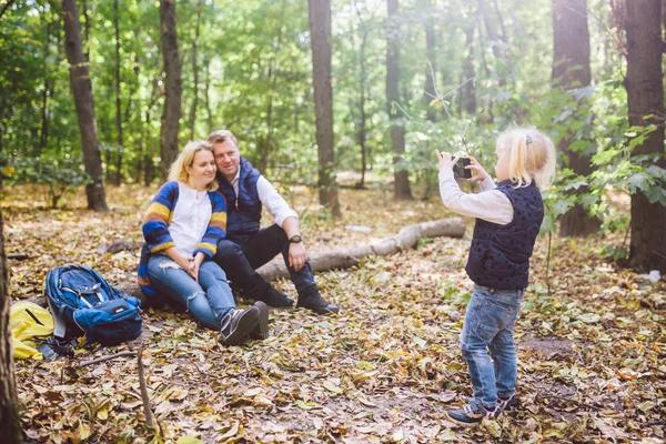 travel, tourism, hike, technology and family concept. Happy child makes photo parents in forest. Mom and dad pose for photo, daughter takes photo on phone. Young family of tourists in wooded area