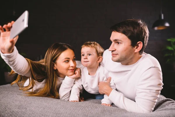A young family lies on the bed and looks at the mobile phone. Mom, dad and son are watching a video on a smartphone in the bedroom in the evening. Family evening vacation at home — Stock Photo, Image