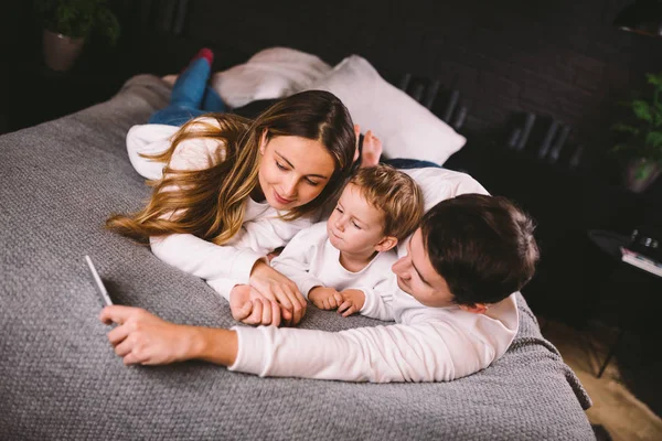 A young family lies on the bed and looks at the mobile phone. Mom, dad and son are watching a video on a smartphone in the bedroom in the evening. Family evening vacation at home