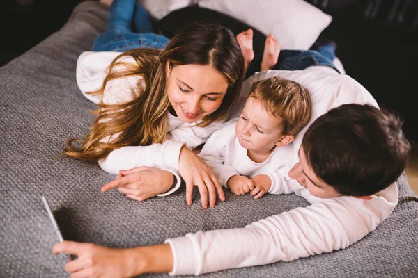 En ung familj ligger på sängen och tittar på mobilen. Mamma, pappa och son tittar på en video på en smartphone i sovrummet på kvällen. Familjekväll semester hemma — Stockfoto