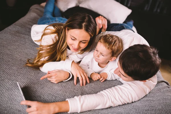 En ung familj ligger på sängen och tittar på mobilen. Mamma, pappa och son tittar på en video på en smartphone i sovrummet på kvällen. Familjekväll semester hemma — Stockfoto