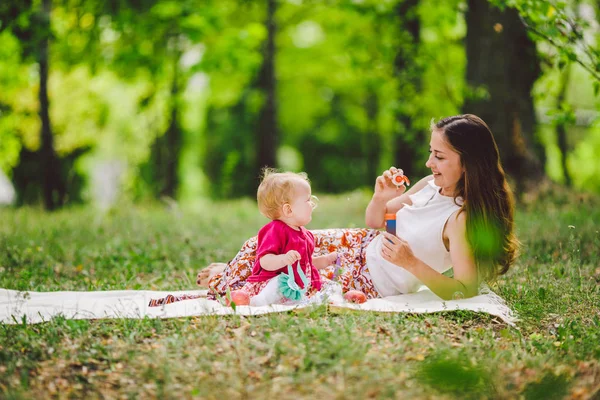 Mujer sonriente jugar y sentarse en la hierba verde en el parque, el descanso y el abrazo mantenga jabón soplador de burbujas con niña linda bebé. Feliz madre e hija en el parque. Recreación familiar al aire libre — Foto de Stock