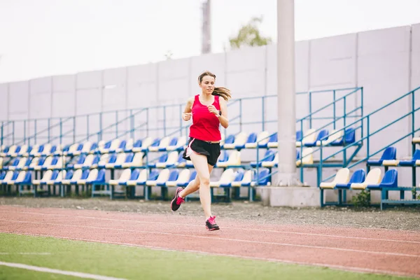 Beautiful young athlete Caucasian woman with big breasts in red T-shirt and  short shorts jogging, running in the stadium with red rubber coating Stock  Photo