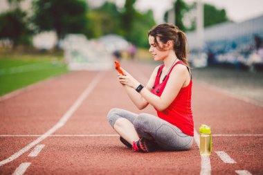 Güzel fitness kızı antrenmandan sonra stadyumda akıllı telefon kullanır. Spor ve sağlıklı. Spor kompleksi içinde kadın cep telefonu kullanımı. Spor ve teknoloji. Çalışmadan sonra dinleniyor, kırmızı yolda koşuyor.