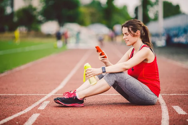 运动后，漂亮的健身女孩在体育馆里用智能手机。 运动与健康。 运动妇女在运动综合体内使用手机。 体育和技术。 运动后休息，在红道上跑 — 图库照片