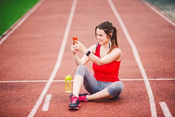 运动后，漂亮的健身女孩在体育馆里用智能手机。 运动与健康。 运动妇女在运动综合体内使用手机。 体育和技术。 运动后休息，在红道上跑 — 图库照片