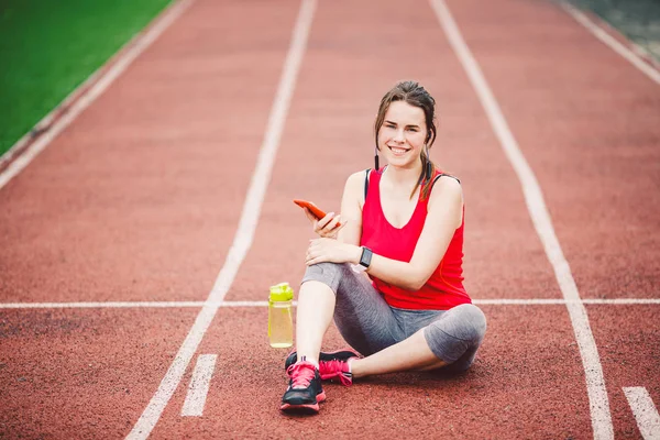 Krásná fitness dívka používá smartphone na stadionu po cvičení. Sport a zdraví. Sportovní žena používá mobil uvnitř sportovního komplexu. Sport a technika. odpočinek po tréninku, běh na červené trati — Stock fotografie