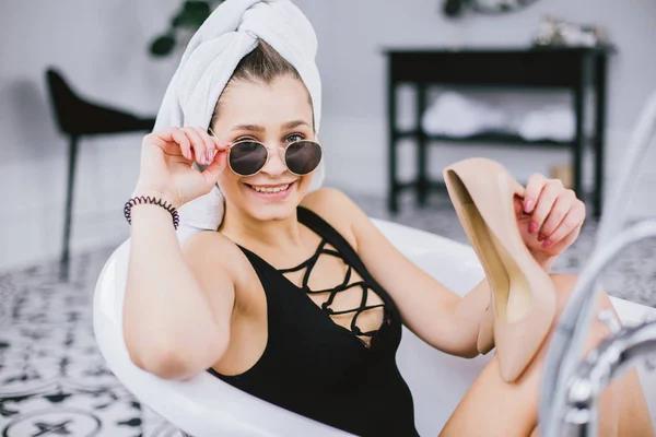 Fashion shot of shopping, young caucasian woman in sunglasses and towel on head holding high heels shoes in hand, lying in bathroom at hotel near window — Stock Photo, Image