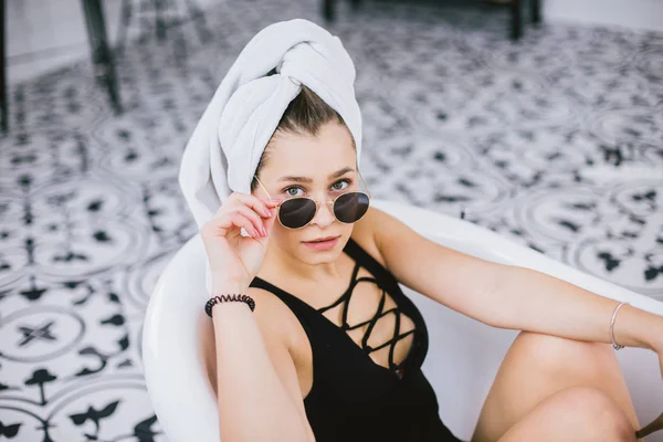 Fashion shot of shopping, young caucasian woman in sunglasses and towel on head holding high heels shoes in hand, lying in bathroom at hotel near window — Stock Photo, Image