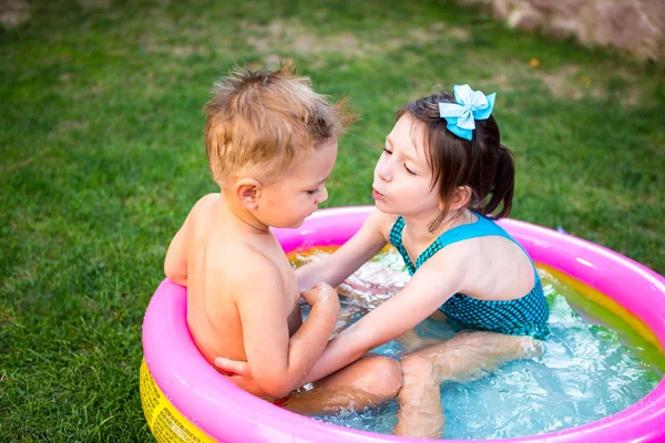 Två lillebror och syster leker och plaskar i poolen den varma sommardagen. Barn simmar i barnpoolen. Två glada söta barn leker och har kul, stänk i uppblåsbar pool på bakgården — Stockfoto