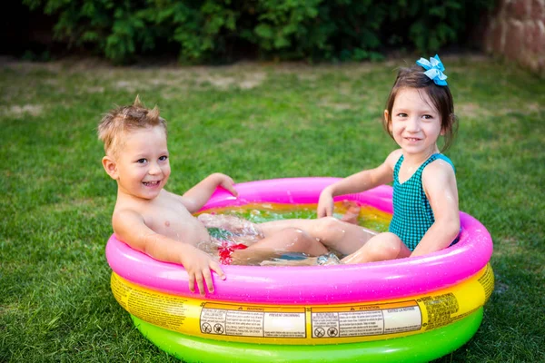 Zwei kleine Geschwister spielen und planschen an heißen Sommertagen im Pool. Kinder schwimmen im Kinderbecken. zwei fröhliche süße Kinder spielen und Spaß haben, planschen im aufblasbaren Pool im Hinterhof — Stockfoto