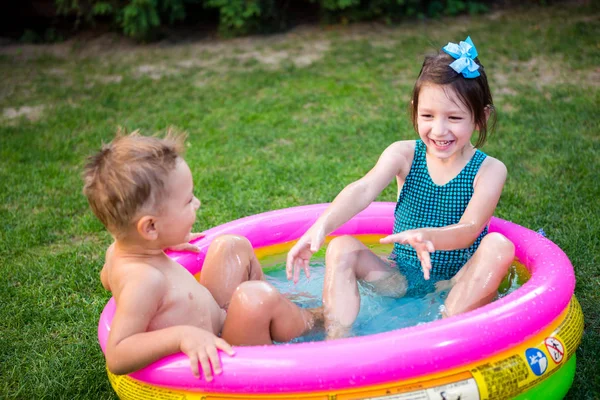 暑い夏の日にプールで遊んでいる2人の弟と妹。子供たちはプールで泳いでいます。2人の陽気なかわいい子供たちが遊んで楽しんで、裏庭でインフレータブルプールでスプラッシュ — ストック写真