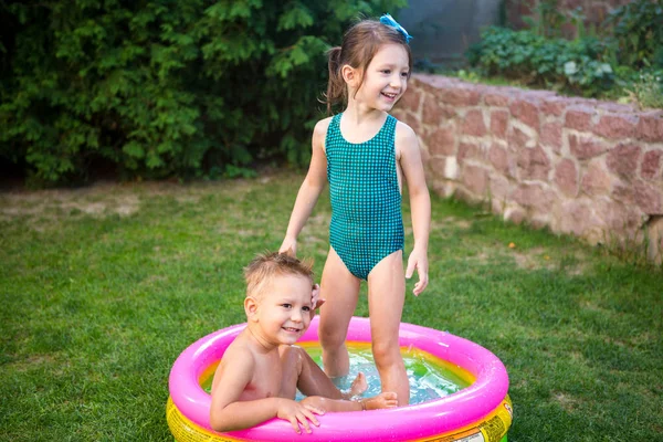 Two little brother and sister playing and splashing in pool on hot summer day. Children swimming in kid pool. Two cheerful cute children playing and having fun, splash in inflatable pool at backyard — Stock Photo, Image