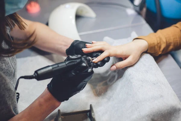 Närbild professionell kosmetolog händer arbetar med elektrisk borrfil på klientfingernaglar. Förfarande tillämpa artificiella naglar. Händerna manikyrist med verktyg. Manicurist handskar tar bort nagelband — Stockfoto