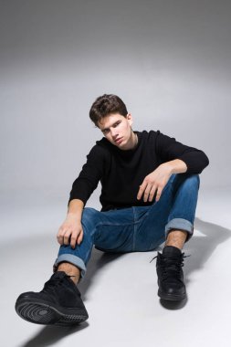 Stylish guy posing in studio sitting on the floor in denim pants and black sweatshirt. Male student portrait full on white background. Studio shot Handsome attractive european man sitting on floor