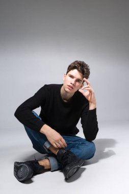 Stylish guy posing in studio sitting on the floor in denim pants and black sweatshirt. Male student portrait full on white background. Studio shot Handsome attractive european man sitting on floor