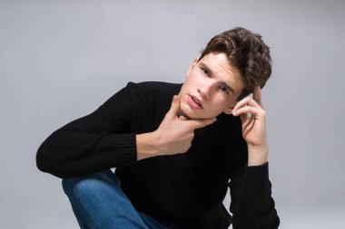 Stylish guy posing in studio sitting on the floor in denim pants and black sweatshirt. Male student portrait full on white background. Studio shot Handsome attractive european man sitting on floor