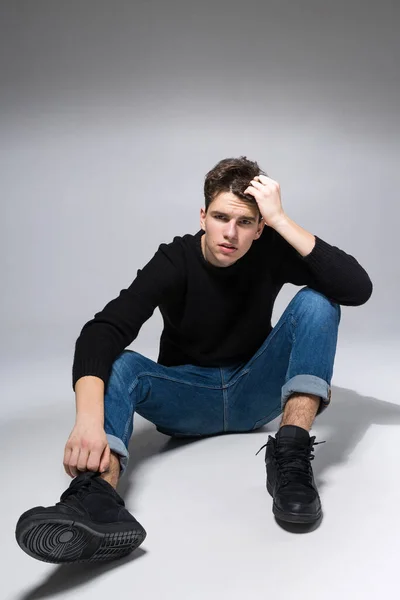 Stylish handsome man posing sitting on floor. Handsome Guy. cool fashion male model sitting on grey background and looking at the camera. Student in a studio in jeans and a black sweater — Stock Photo, Image