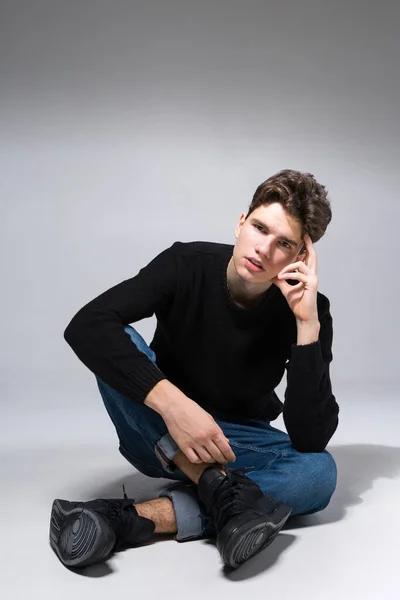 Elegante bell'uomo che posa seduto sul pavimento. Un bel ragazzo. cool moda modello maschile seduto su sfondo grigio e guardando la fotocamera. Studente in uno studio in jeans e maglione nero — Foto Stock