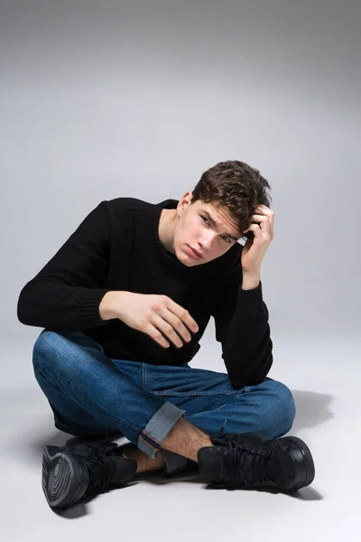 Ragazzo elegante in posa in studio seduto sul pavimento in pantaloni di jeans e felpa nera. Ritratto studentesco maschile completo su sfondo bianco. Studio shot Bello attraente uomo europeo seduto sul pavimento — Foto Stock