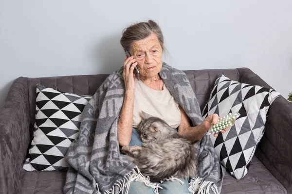 Una anciana enferma tomando medicinas en casa. Mujer mayor hablando por teléfono móvil mientras mira la medicina en el dormitorio. pensionista estudiando medicina llamando doctor mientras sentado en sofá con gato —  Fotos de Stock