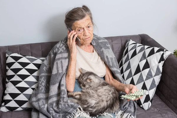 Больная старшая женщина принимает лекарства дома. Старшая женщина разговаривает по мобильному телефону во время просмотра лекарств в спальне. Пенсионер изучает медицину, вызывая врача, сидя на диване с котом — стоковое фото