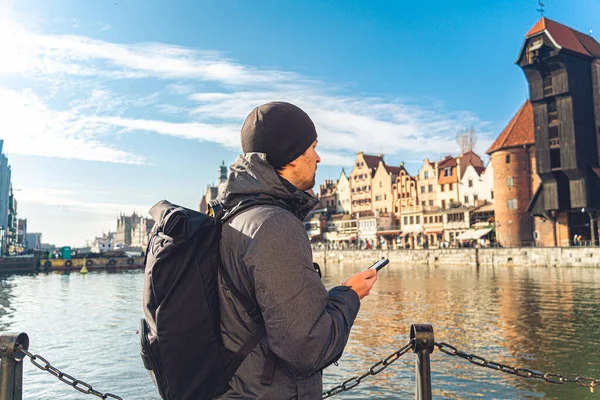 Theme of tourism in Poland. Traveling in Gdansk. A male traveler uses a phone on the embankment of the Motlawa River amid the main attraction, a symbol of the city of Gdansk Gate crane — Zdjęcie stockowe