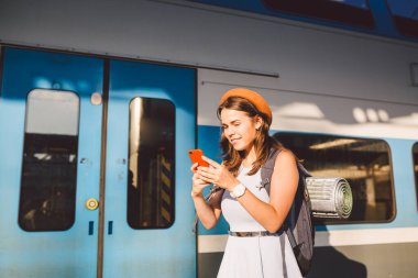 İstasyonda akıllı telefon kullanan gülümseyen kadın. Akıllı telefon tutan ve tren istasyonunda duran genç bir kadın. Tema Taşımacılık ve Seyahat. yolculuk konsepti.
