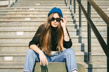 Mavi kot pantolon giymiş, kaykayla poz veren şık giyinmiş bir kadın. Sokak fotoğrafı. Kaykay tutan kızın portresi. Yaşam tarzı, gençlik kavramı. Boş zaman, hobi ve şehirde paten..