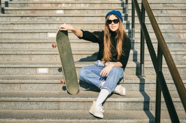 Mavi kot pantolon giymiş, kaykayla poz veren şık giyinmiş bir kadın. Sokak fotoğrafı. Kaykay tutan kızın portresi. Yaşam tarzı, gençlik kavramı. Boş zaman, hobi ve şehirde paten..