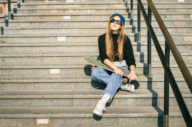 Mavi kot pantolon giymiş, kaykayla poz veren şık giyinmiş bir kadın. Sokak fotoğrafı. Kaykay tutan kızın portresi. Yaşam tarzı, gençlik kavramı. Boş zaman, hobi ve şehirde paten..