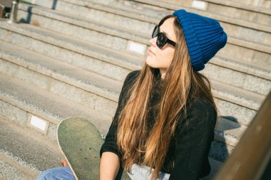 Mavi kot pantolon giymiş, kaykayla poz veren şık giyinmiş bir kadın. Sokak fotoğrafı. Kaykay tutan kızın portresi. Yaşam tarzı, gençlik kavramı. Boş zaman, hobi ve şehirde paten..