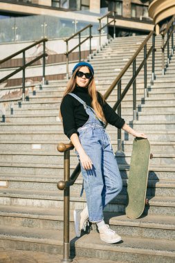 Sokakta elinde kaykayla poz veren beyaz bir kadın. Şehir ortamında kot pantolon giyen genç bir kız ekstrem spor yapıyor. Gençlik eğlencesi teması, yaşam tarzı. Şehirde kaykaycı..