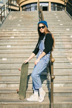 Mavi kot pantolon giymiş, kaykayla poz veren şık giyinmiş bir kadın. Sokak fotoğrafı. Kaykay tutan kızın portresi. Yaşam tarzı, gençlik kavramı. Boş zaman, hobi ve şehirde paten..