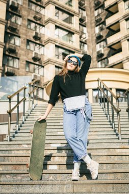 Mavi kot pantolon giymiş, kaykayla poz veren şık giyinmiş bir kadın. Sokak fotoğrafı. Kaykay tutan kızın portresi. Yaşam tarzı, gençlik kavramı. Boş zaman, hobi ve şehirde paten..