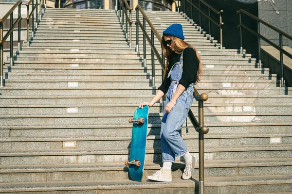 スケートボードとポーズをとるブルーデニムジャンプスーツのスタイリッシュな服を着た女性 ストリートフォト スケートボードを保持する少女の肖像画 ライフスタイル 若者のコンセプト 市内のレジャー スケート — ストック写真