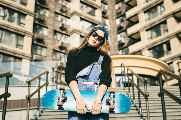 スケートボードとポーズをとるブルーデニムジャンプスーツのスタイリッシュな服を着た女性 ストリートフォト スケートボードを保持する少女の肖像画 ライフスタイル 若者のコンセプト 市内のレジャー スケート — ストック写真