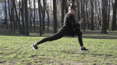 Parkta spor yaparken cep telefonu kullanan çekici bir kadın. Sağlıklı genç bir kadın dışarıda ısınıyor. Akıllı telefondaki sosyal ağlardaki yazışmalar dikkatini dağıtıyor. sağlıklı fitness yaşam tarzı